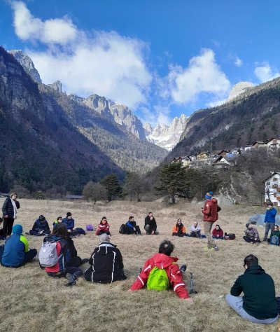 un gruppo di persone sedute per terra, in cerchio, in un prato montano. sullo sfondo, alcune cime delle Dolomiti