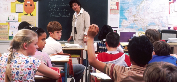 Aula scolastica con insegnante alla lavagna e bambini che la ascoltano e attendono di poter fare domande con la mano alzata