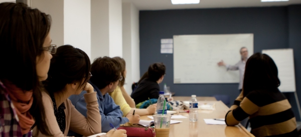 gruppo di insegnanti, attorno ad un tavolo, che ascolta la lezione di un professore che è accanto ad una lavagna