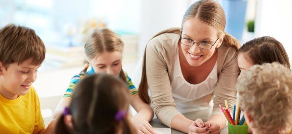 maestra bionda china su un tavolo attorno al quale sono seduti cinque bambini