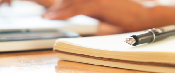 una penna sopra a dei fogli e due mani sullo sfondo che prendono altri fogli