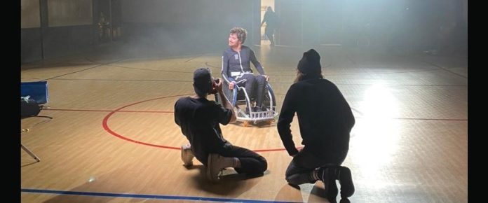 Michele in una palestra seduto in carrozzina emhra indossa la tuta Exopluse e viene fotografato da due persone