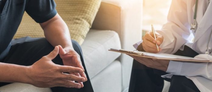 dettagli delle mani di due uomini seduti uno di fronte all'altro, uno dei quali indossa un camice, porta uno stetoscopio al collo e scrive su una cartellina