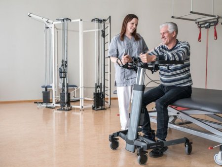 uomo si aiuta con il deambulatore per alzarsi dal letto sul quale è seduto. vicino a lui, una operatrice in piedi