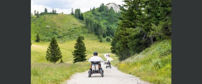 alcune persone lungo una strada circondata di montagne, scengono utilizzando il quad per disabili Zoom